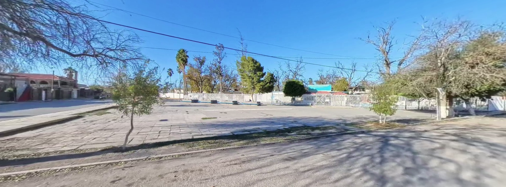 Fotografía en la que se muestra una explanada. Al fondo hay una reja de color blanco que rodea las instalaciones y en medio de la reja se encuentra el acceso al plantel.